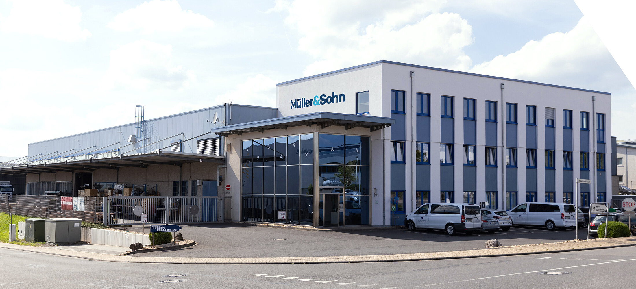 Moderne Firmenzentrale von Müller & Sohn mit Bürogebäude und angrenzender Lager- oder Produktionshalle, davor geparkte Firmenfahrzeuge